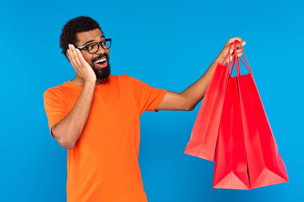 zdumiony Afrykański Amerykanin w okularach trzyma torby na zakupy odizolowane na niebiesko  - Zdjęcie, obraz