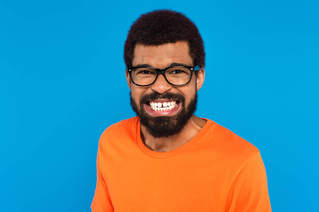 stresszes afro-amerikai férfi szemüveges mutatja fehér fogak elszigetelt kék - Fotó, kép