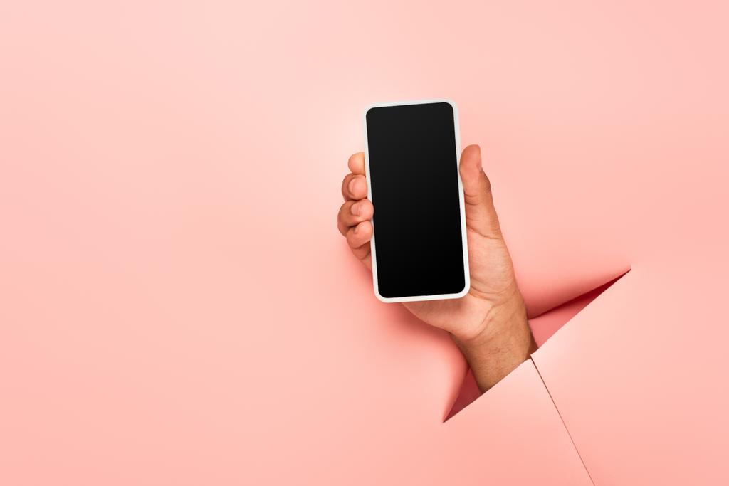 partial view of african american man holding smartphone with blank screen on ripped pink background  - Photo, Image