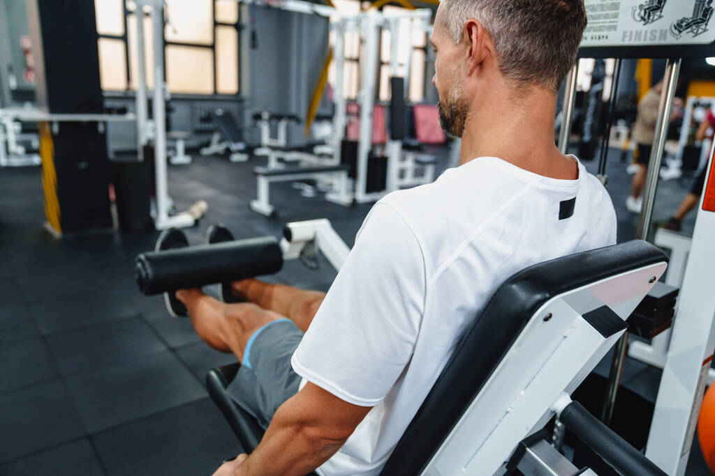 attrayant confiant homme sportif sain en utilisant la machine de boucle de jambe assise dans la salle de gym, vue latérale - Photo, image