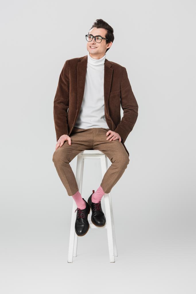 счастливый человек в винтажной одежде смотрит в сторону, сидя на высоком стуле на сером фоне - Фото, изображение