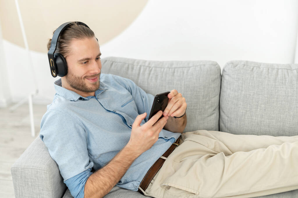 Ικανοποιημένος όμορφος άντρας που χρησιμοποιεί smartphone και ασύρματα ακουστικά για να ακούει μουσική - Φωτογραφία, εικόνα