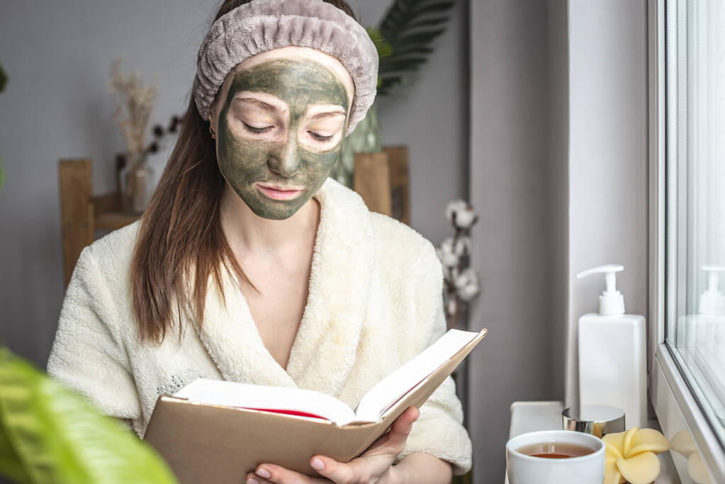 Kaunis nuori nainen kylpytakissa ja vihreä kosmeettinen naamio kasvoillaan lukee kirjaa ja juo teetä. Rentoutumisen, kauneushoitojen, ihonhoidon ja elämäntavan käsite - Valokuva, kuva