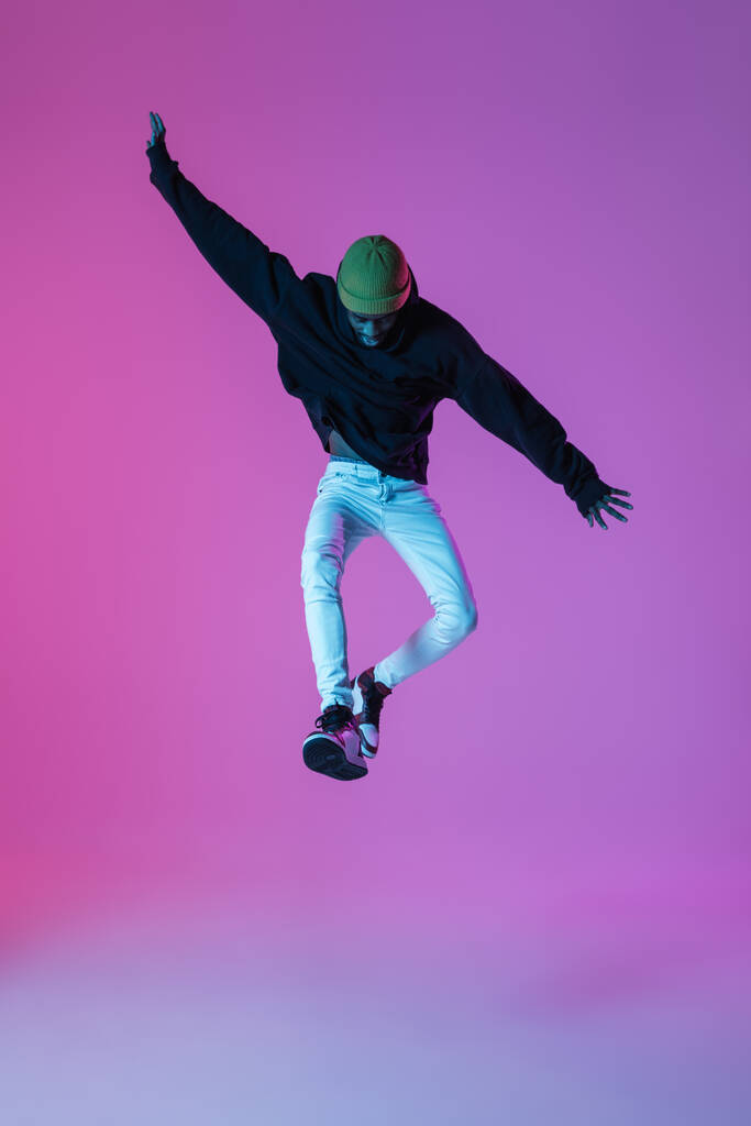 Young stylish man in modern street style outfit isolated on gradient background in neon light. African-american fashionable model in look book, musician performing. - Photo, Image