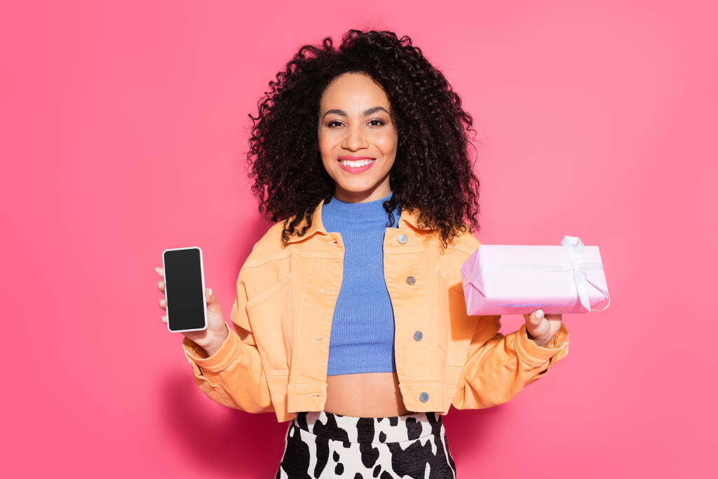 curly african american woman in crop top and jacket holding smartphone with blank screen and present on pink  - Photo, Image