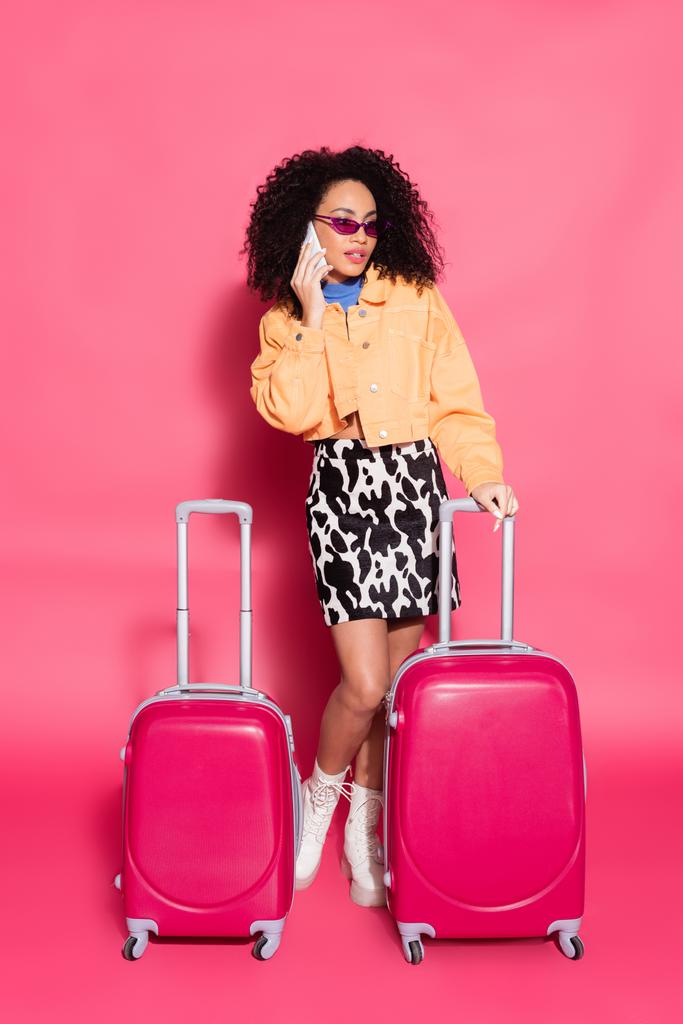 ピンクのスーツケースの近くでスマートフォンで話しているアフリカ系アメリカ人女性の  - 写真・画像