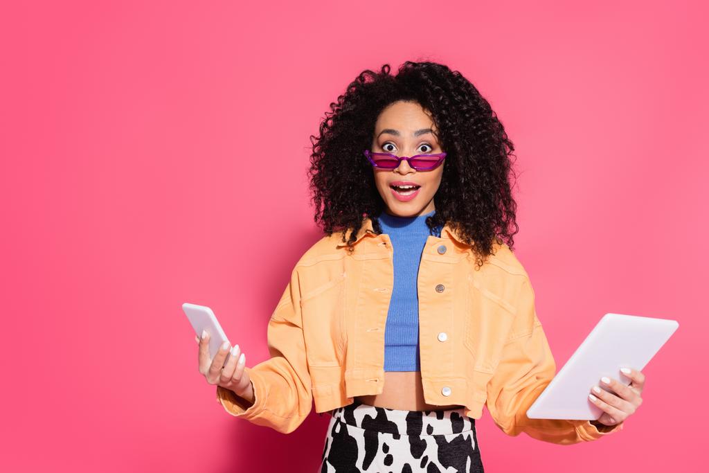 zavaros afro-amerikai nő napszemüvegben tartó okostelefon és digitális tabletta rózsaszín - Fotó, kép