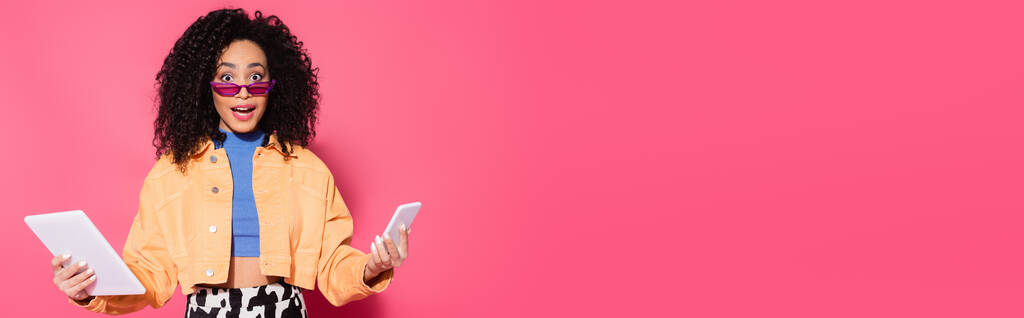 mujer afroamericana confusa en gafas de sol sosteniendo teléfono inteligente y tableta digital en rosa, bandera - Foto, imagen