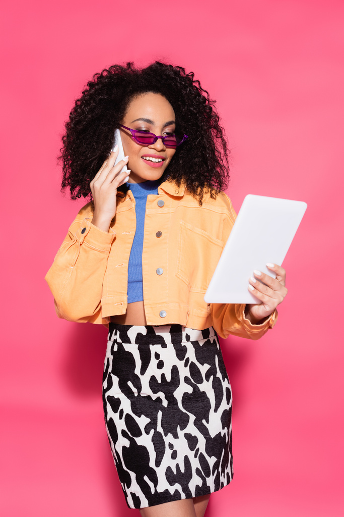 felice donna afroamericana in occhiali da sole che parla sullo smartphone mentre tiene il tablet digitale su rosa - Foto, immagini