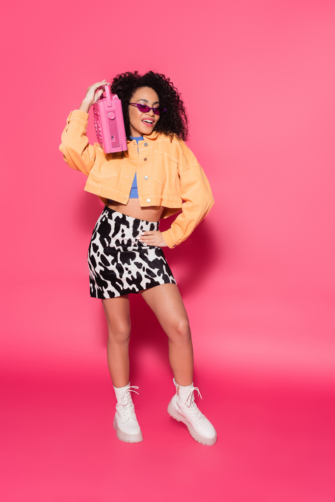 piena lunghezza di felice donna afro-americana in occhiali da sole in possesso di boombox e in posa con mano sul fianco su rosa  - Foto, immagini