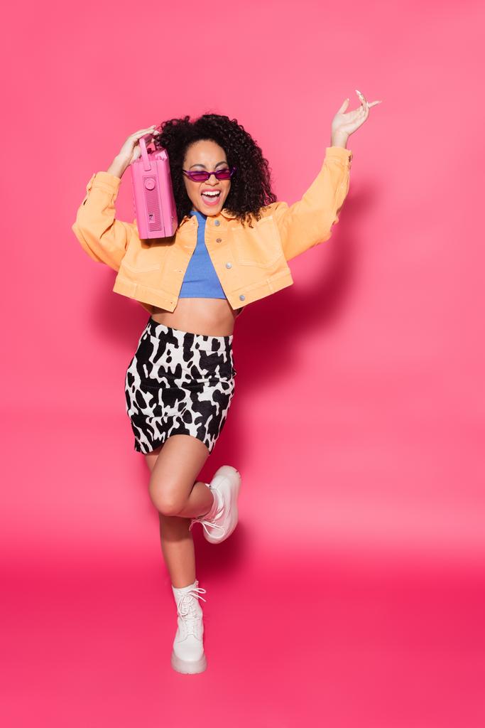 piena lunghezza di felice donna africana americana in occhiali da sole in possesso di boombox e in posa su rosa  - Foto, immagini