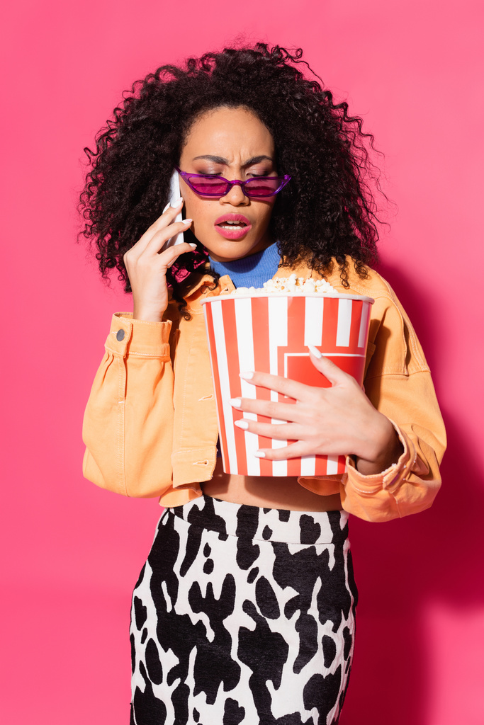 afro-amerikai nő napszemüvegben kezében egy vödör popcorn és beszél okostelefonon rózsaszín  - Fotó, kép