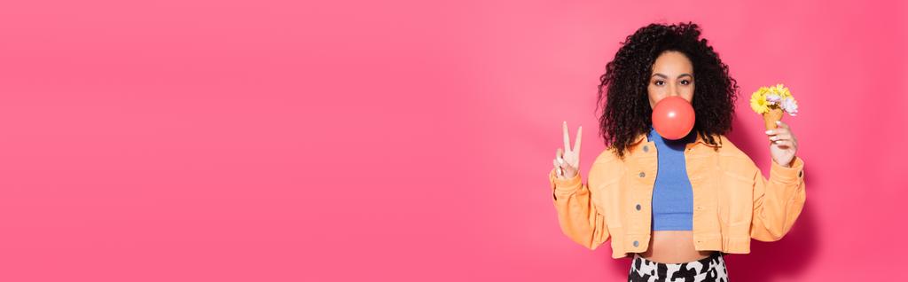curly african american woman blowing bubble gum, holding waffle cone with flowers and showing peace sign on pink, banner - Photo, Image