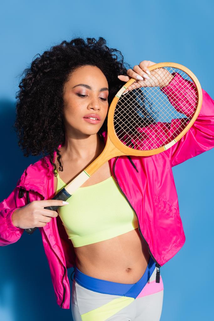 curly african american woman in crop top posing with tennis racket on blue - Photo, Image