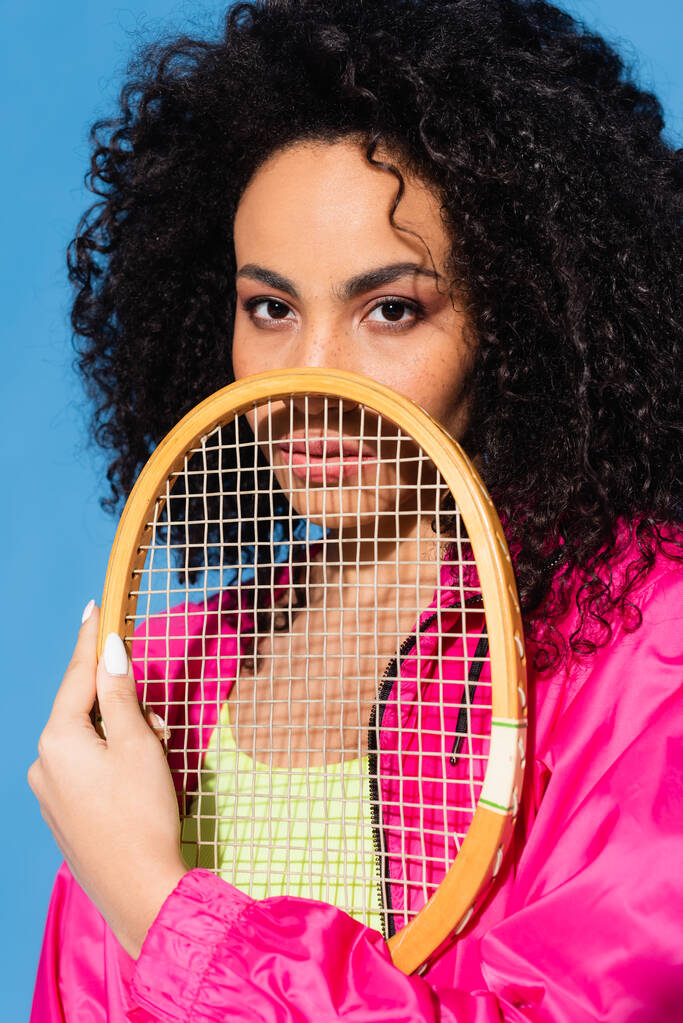 кучерява афроамериканська жінка позує з тенісною ракеткою, дивлячись на камеру ізольовано на синьому
 - Фото, зображення
