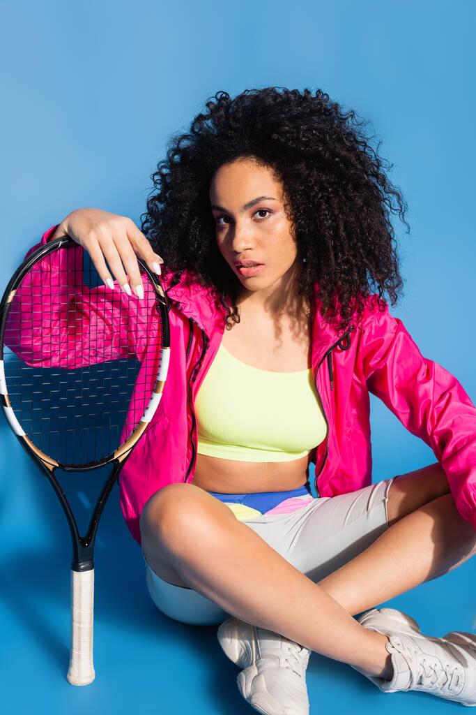 Αφροαμερικανή που κρατάει ρακέτα του τένις και κάθεται σε μπλε χρώμα. - Φωτογραφία, εικόνα
