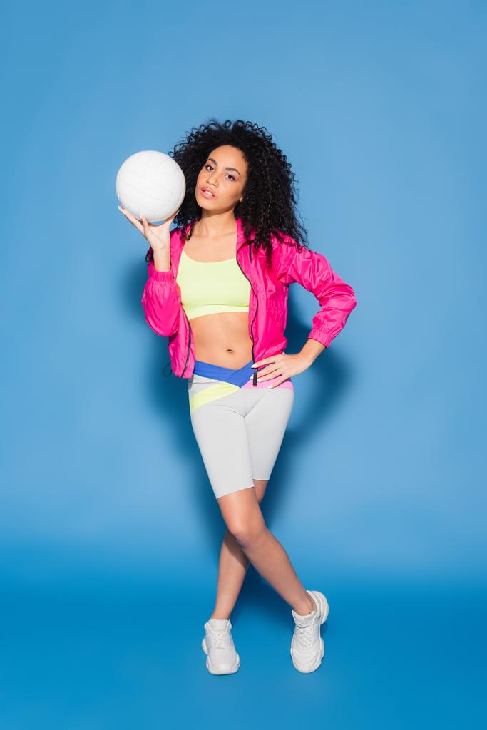 full length of sporty african american woman in pink jacket holding volleyball while posing with hand on hip on blue - Photo, Image