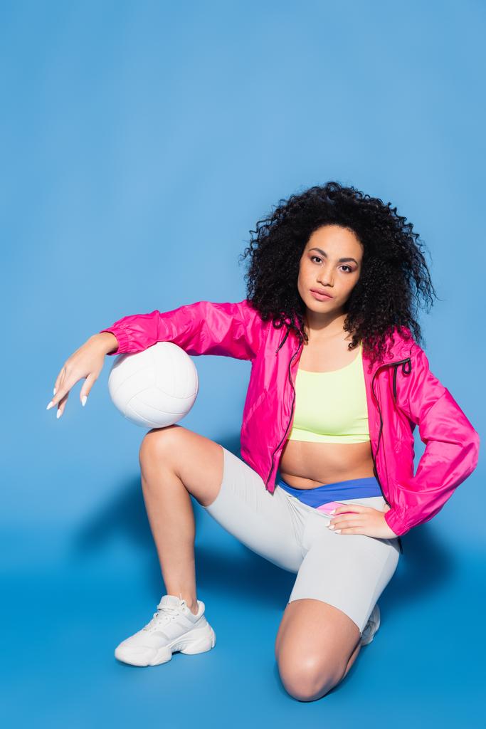 young african american woman in pink jacket sitting with volleyball on blue - Photo, Image