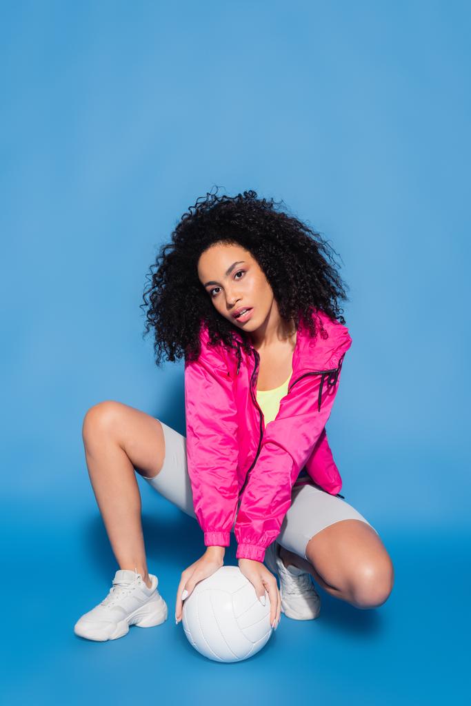 giovane donna afroamericana in giacca rosa in posa vicino pallavolo su blu - Foto, immagini