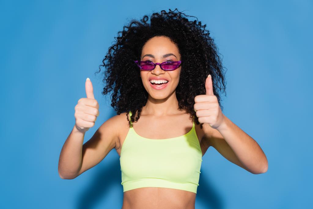 Χαρούμενη Αφροαμερικανή με γυαλιά ηλίου που δείχνει τους αντίχειρες στο μπλε. - Φωτογραφία, εικόνα