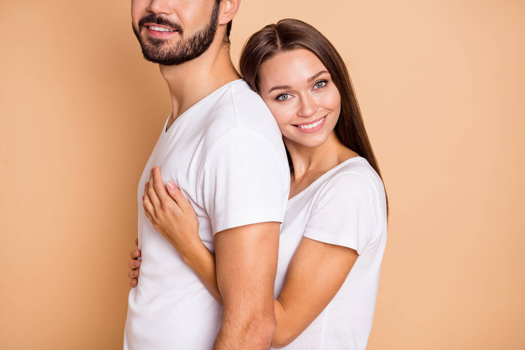 Περικοπή φωτογραφία του γοητευτικό λαμπερό παντρεμένο ζευγάρι ντυμένο λευκά t-shirts αγκαλιάζει πίσω χαμογελώντας απομονωμένο φόντο μπεζ χρώμα - Φωτογραφία, εικόνα