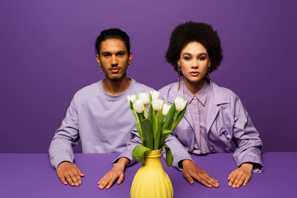 couple afro-américain assis près du vase avec des tulipes blanches isolées sur violet - Photo, image