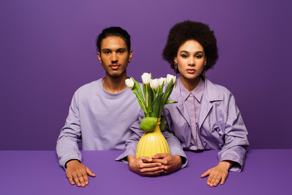 à la mode jeune couple afro-américain assis près du vase avec des tulipes isolées sur violet - Photo, image