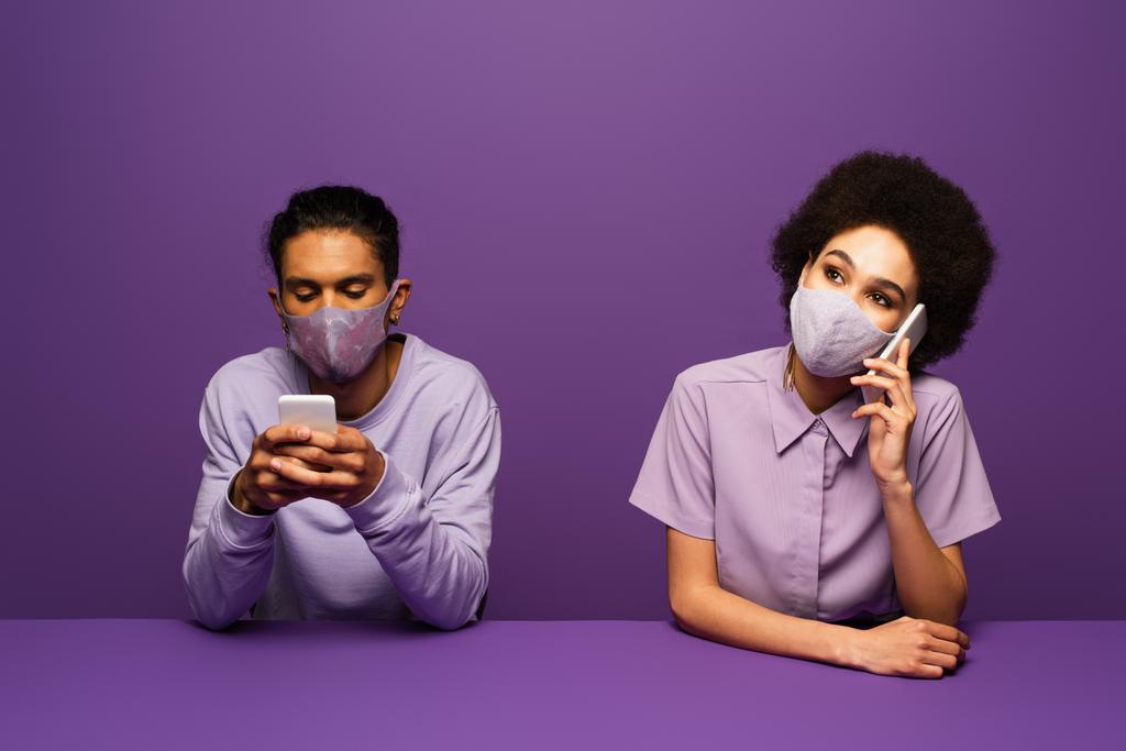 молодий афроамериканський чоловік і жінка в захисних масках, використовуючи смартфони ізольовані на фіолетовому
  - Фото, зображення
