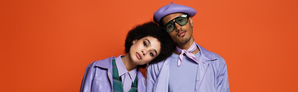 curly woman in purple jacket leaning on model in beret and sunglasses isolated on orange, banner - Photo, Image