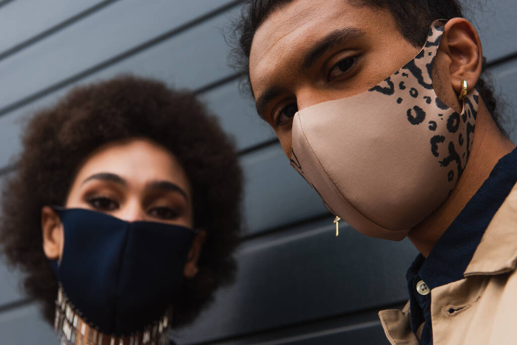 молодой африканский американец и размытая женщина в стильных защитных масках, позирующих снаружи  - Фото, изображение