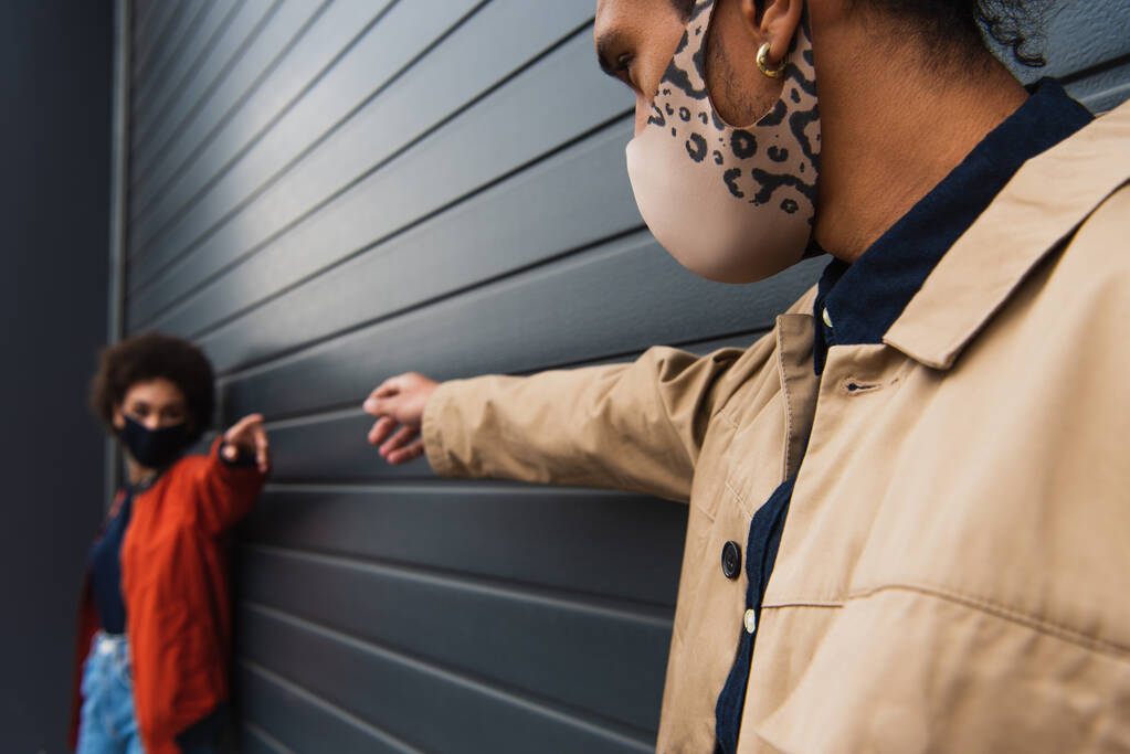 молодий афроамериканський чоловік і розмита жінка в стильних захисних масках з розтягнутими руками
  - Фото, зображення