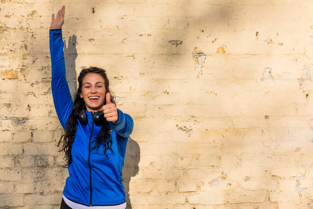 Portret szczęśliwej sportowej kobiety pokazującej kciuk w górę stojąc przed ścianą . - Zdjęcie, obraz