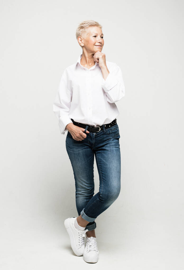 Pełna długość zdjęcie stylowej kobiety w dżinsach i białej bluzce z krótkimi białymi włosami pozowanie na jasnym szarym tle. - Zdjęcie, obraz