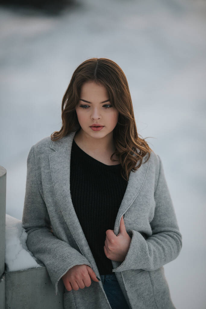 A vertical shot of a sexy woman with a grey jacket leaning on the wall on a snowy park - Photo, Image