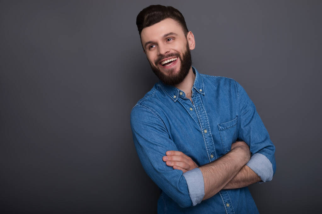 Щасливий збуджений молодий бородатий чоловік в джинсах з схрещеними руками позує на сірому фоні
 - Фото, зображення
