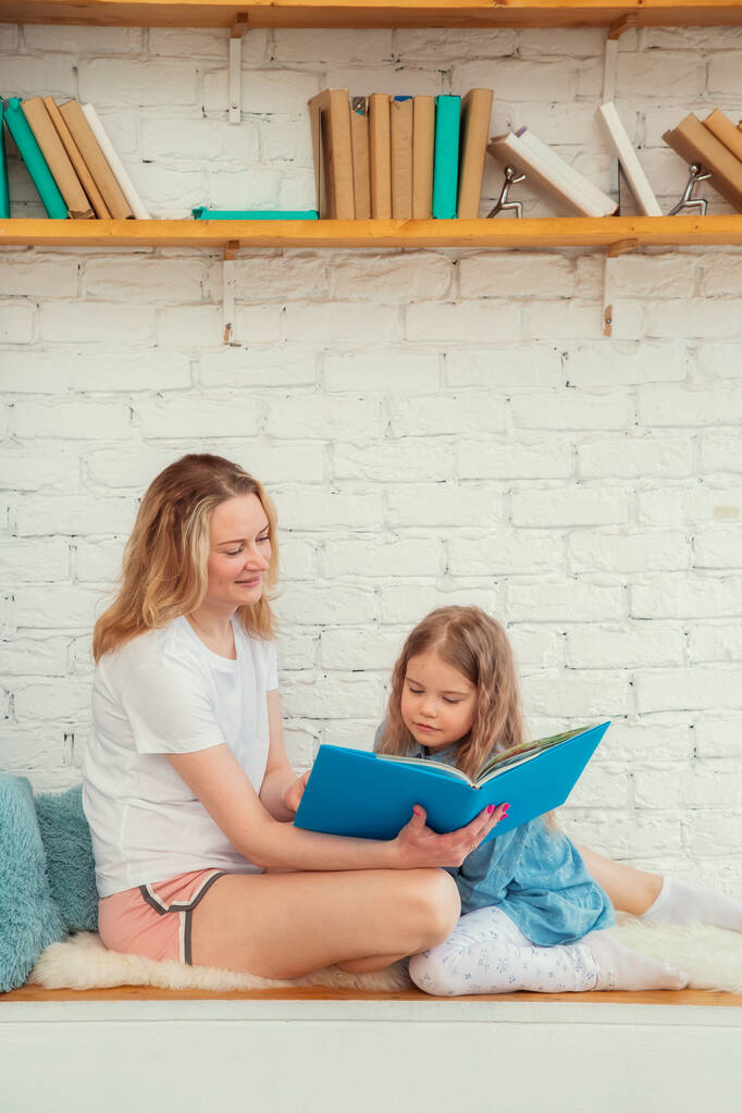 Feliz madre caucásica con su hija leyendo un libro y sonriendo mientras está sentada en el suelo en la sala de estar - Foto, Imagen