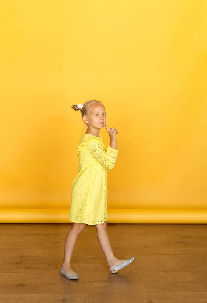 Cute little blonde with bright emotions - attention, mystery, silence, interest. She is wearing a simple yellow dress. Girl`s hand is raised, a sign of silence and attention. Yellow background. European race. - Photo, Image