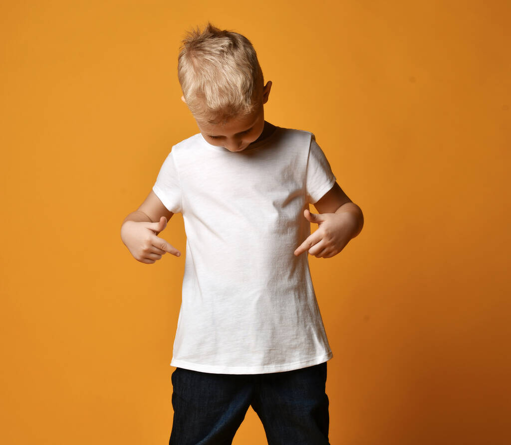 Blonďatý chlapec dítě v bílém tričku a džíny stojící a dívající se na sebe dolů přes žluté pozadí - Fotografie, Obrázek