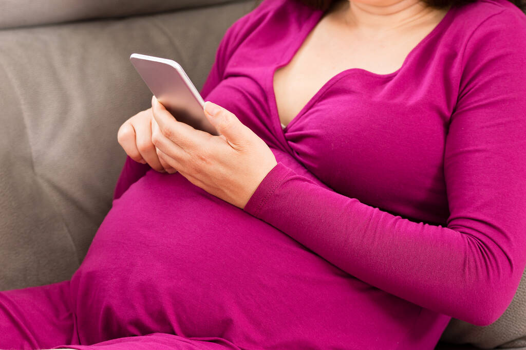 Közelkép egy terhes nőről, aki otthon sms-ezik a mobilján. - Fotó, kép