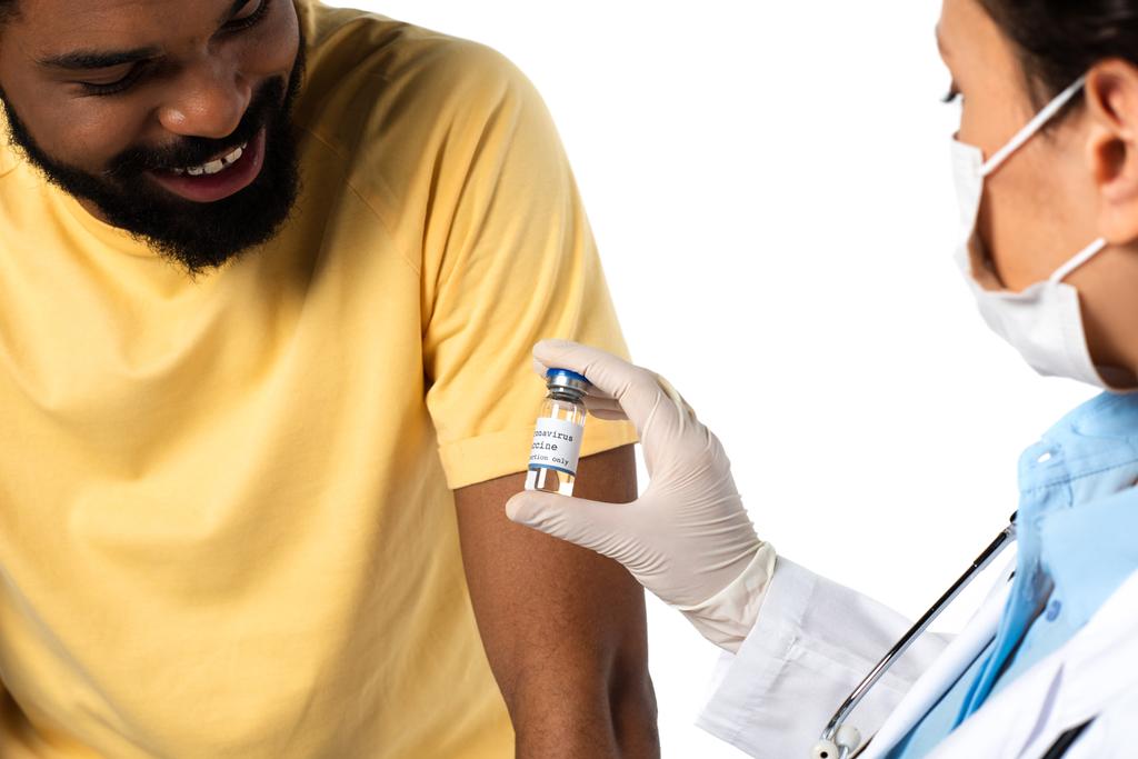 Vaccin en main du médecin au premier plan flou près d'un patient afro-américain souriant isolé sur blanc  - Photo, image