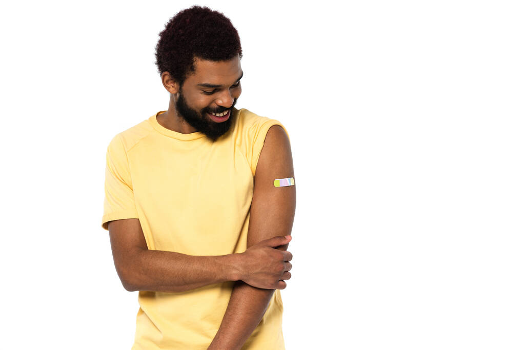 Χαμογελώντας Αφροαμερικάνος κοιτάζοντας αυτοκόλλητο γύψο στο χέρι απομονώνονται σε λευκό  - Φωτογραφία, εικόνα