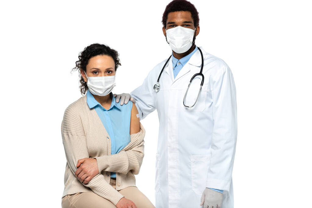 Африканский американский врач в латексных перчатках, стоящий рядом с пациентом в медицинской маске и клей на руке изолирован на белом  - Фото, изображение