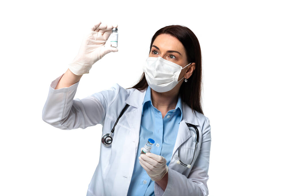 Μελαχρινή γιατρός με ιατρική μάσκα κοιτάζοντας το εμβόλιο που απομονώνεται σε λευκό  - Φωτογραφία, εικόνα