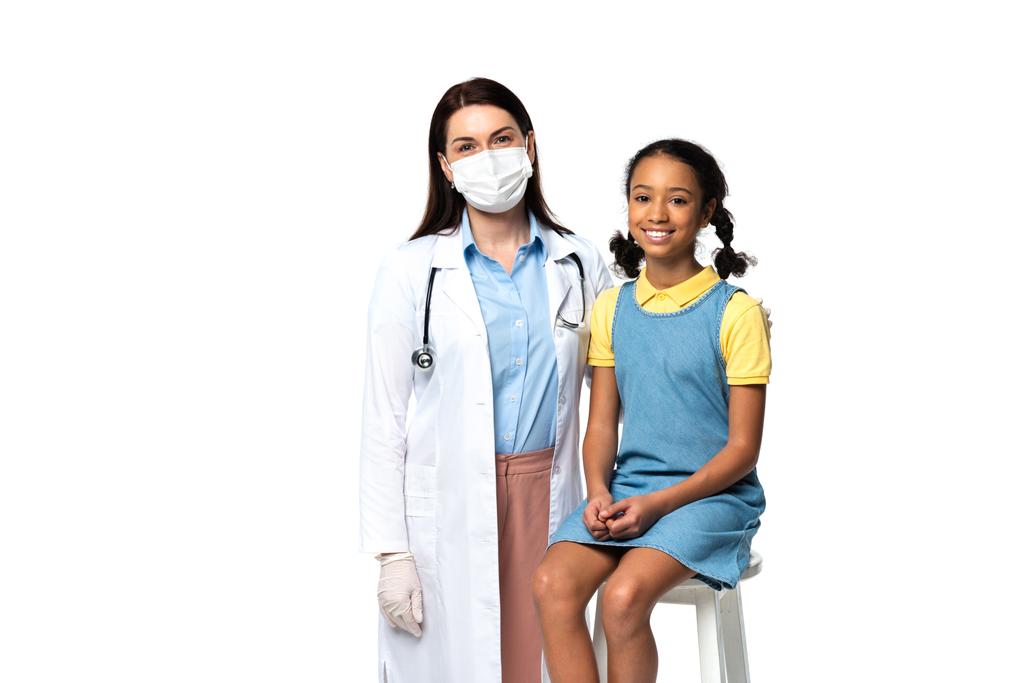 Γιατρός με ιατρική μάσκα που στέκεται κοντά σε χαμογελαστό αφροαμερικανό παιδί απομονωμένο στα λευκά.  - Φωτογραφία, εικόνα