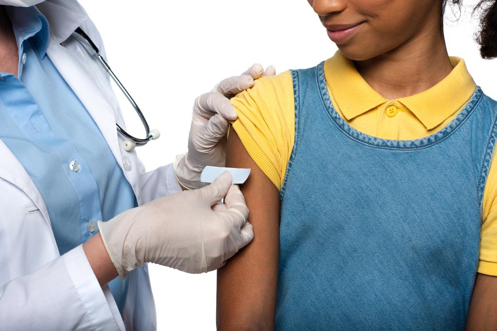 Widok lekarza nakładającego plaster na ramię amerykańskiego dziecka wyizolowanego na biało  - Zdjęcie, obraz