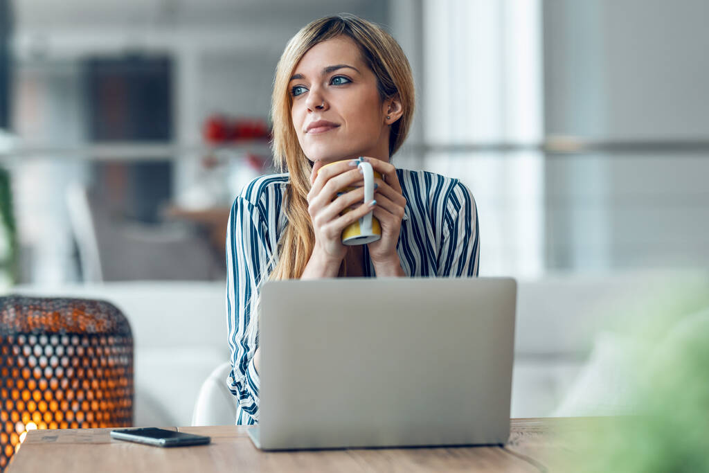 Знімок красивої жінки, що працює з ноутбуком, п'є чашку кави, чекаючи у вітальні вдома
. - Фото, зображення