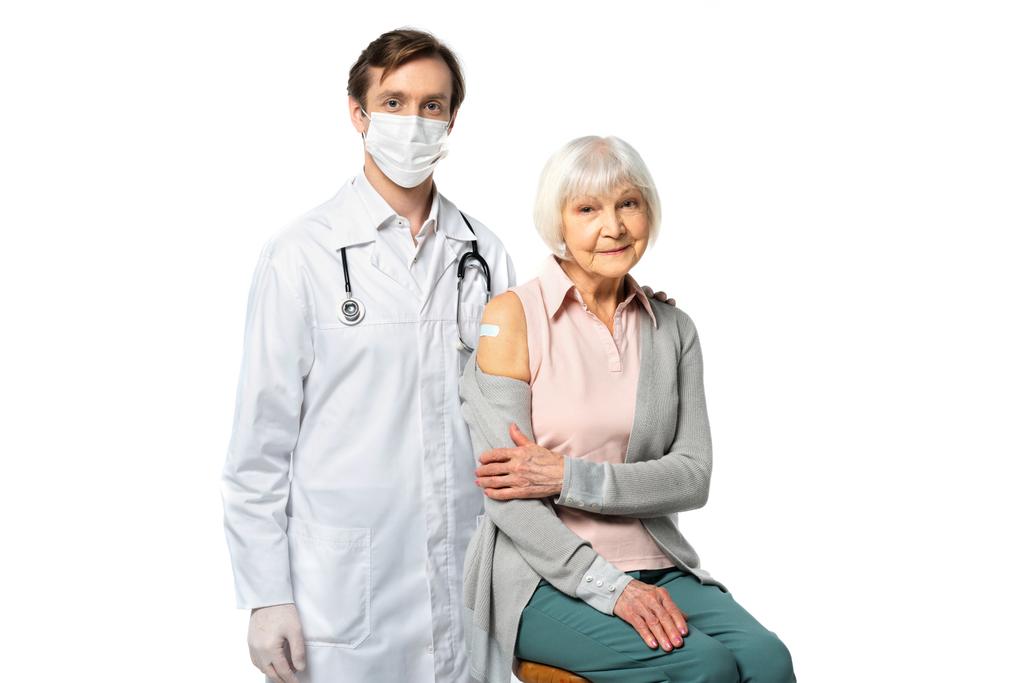 Врач в медицинской маске обнимает пожилого пациента клеевой штукатуркой, изолированной на белом  - Фото, изображение