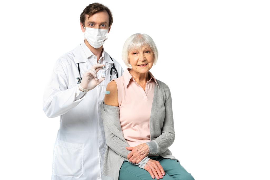 Lékař v lékařské masce drží vakcínu v blízkosti usmívající se pacient s lepicí cestou izolované na bílém  - Fotografie, Obrázek