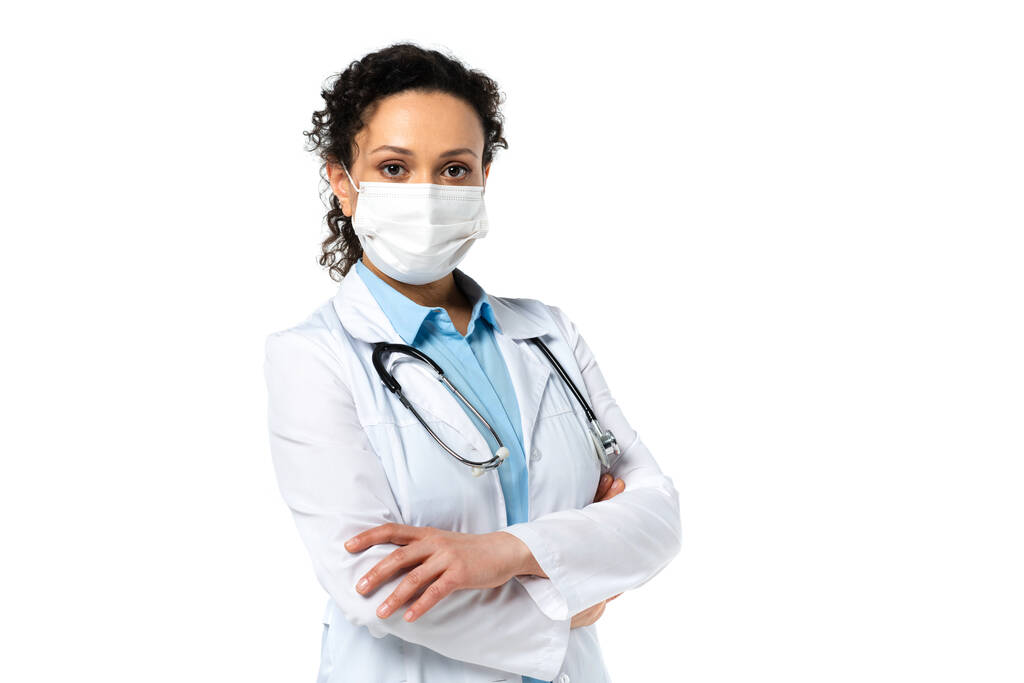 Médico afroamericano en máscara médica mirando a la cámara mientras está de pie aislado en blanco  - Foto, imagen