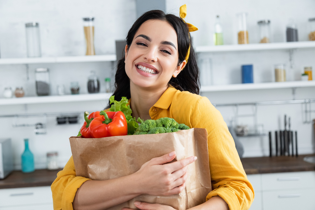 ευτυχισμένη μελαχρινή γυναίκα αγκαλιάζει χάρτινη τσάντα με φρέσκα λαχανικά  - Φωτογραφία, εικόνα